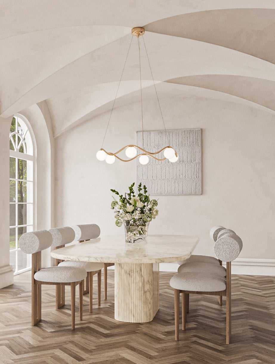 This pendant lamp in neutral-colored interior with a marble table and arches on the ceiling. The chandelier collection features sculptural, wave-inspired brass frames with spherical frosted glass globes, adding fluid motion and sophistication to